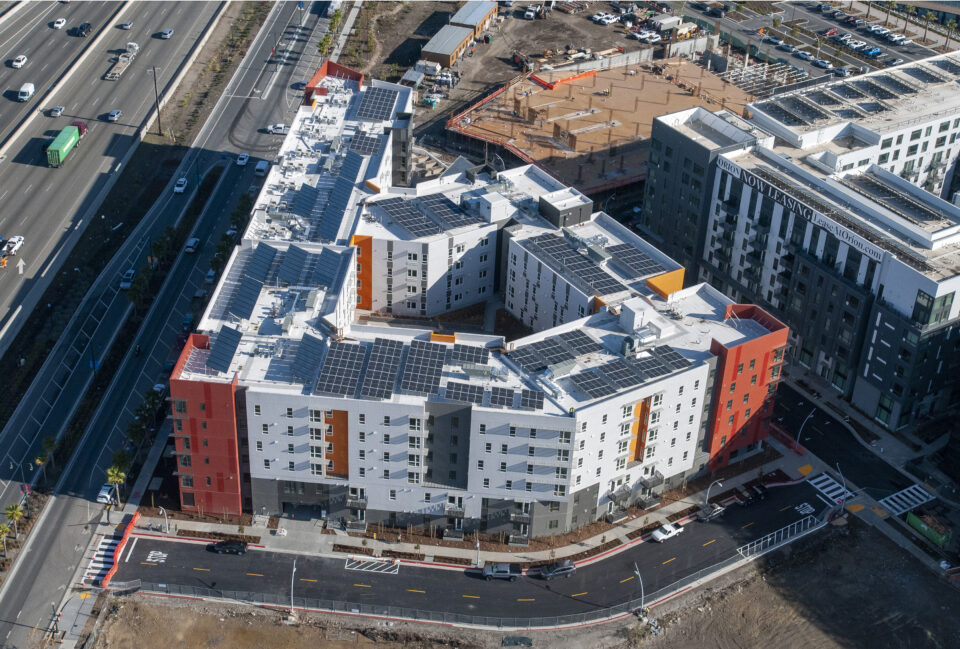 Image of a multi-family affordable apartment home from the sky - white and red sides, solar panels on top, many windows. 