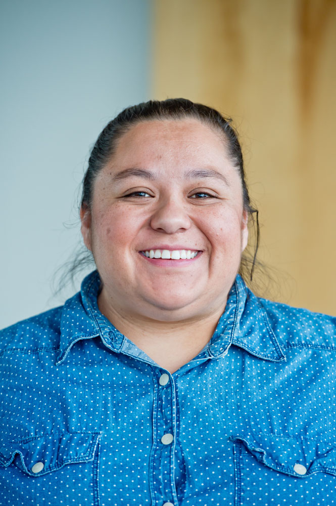 Dolores Tejada smiles at the camera. Dolores is a brown nonbinary person, with hair slicked back into a ponytail. They are wearing a blue button down shirt.