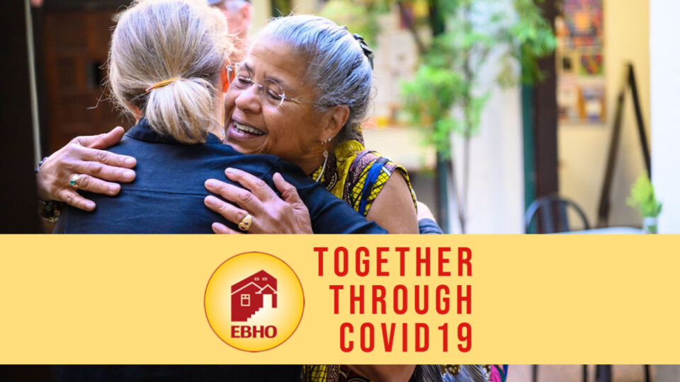 Image of two women hugging underneath a yellow banner that reads "Together through COVID19"