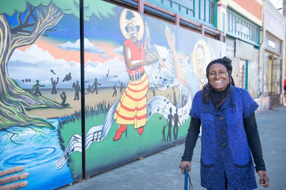 Denise, a Black elder wearing a bright blue top, smiles while looking at the camera in front of a bright mural of Ohlone people.