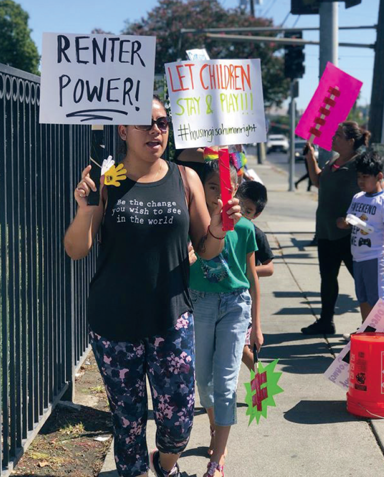 A Latina mother and her child match at a protest in Concord carrying signs that read "Renter Power!" and "Let children stay and play! #HousingIsAHumanRight".