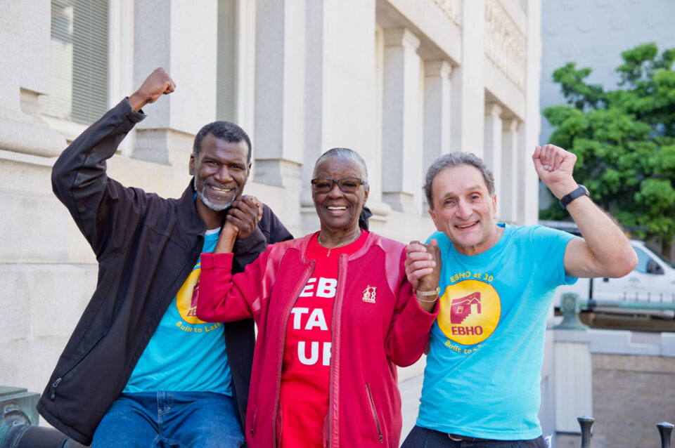 Three residents of affordable housing in EBHO shirts hold hands and raise their fists.
