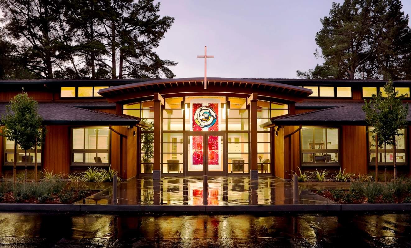 Picture of the Lafayette-Orinda Presbyterian Church on a rainy day at dusk. There is a red stained glass window featuring a rose on the door of the church. You can see the reflection of the window's colors in the puddle in front of the church.