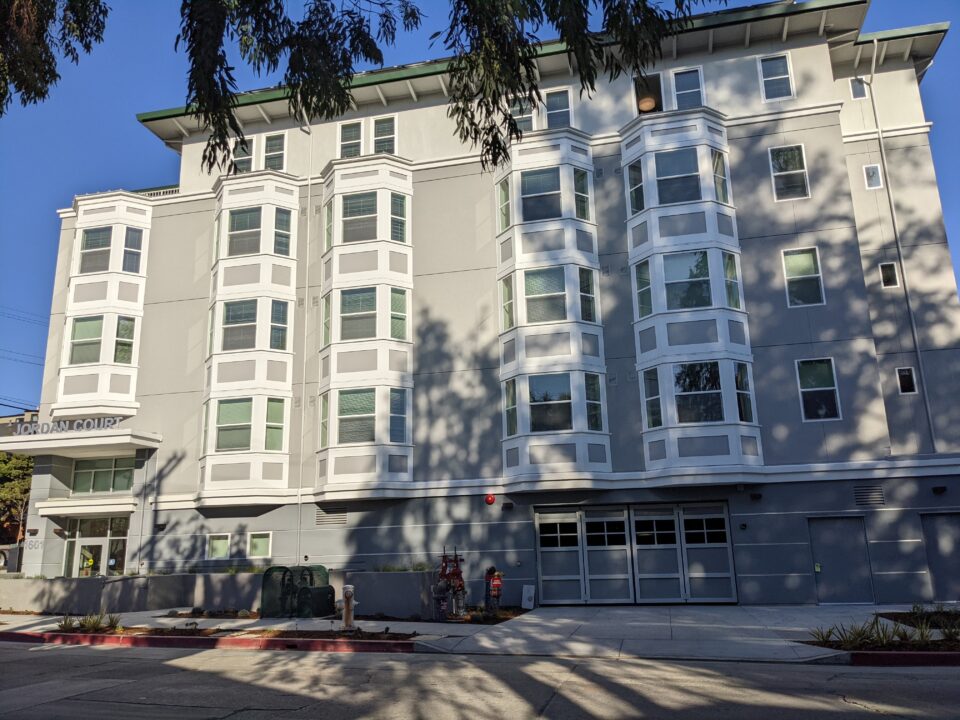 Photo of the Jordan Court affordable housing building for senior citizens in Berkeley. The building is five stories tall and it is grey with classic bay window-style architectural features. An overhang over the front door on the left reads "Jordan Court".