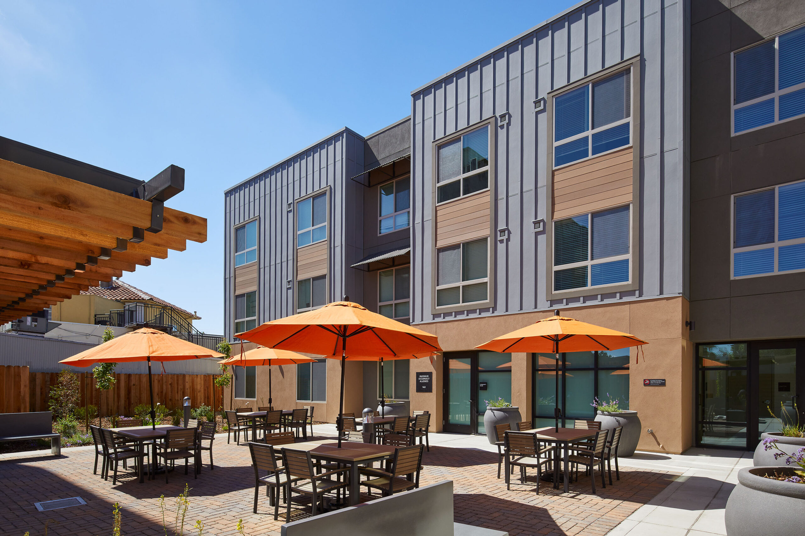 Photo of a sunny courtyard at the Veterans Square development. Orange umbrellas cover dining tables in front of the building.