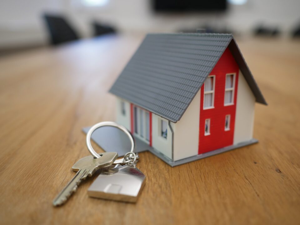 A set of home keys sitting on a wooden table. The keys are placed next to a miniature model of a home painted white with red accents.