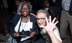 Two members of EBHO's Regional Community Organization Program pose for a photo at EBHO's 2019 Membership Meeting. The man sitting in front has five fingers raised to wave at the camera.