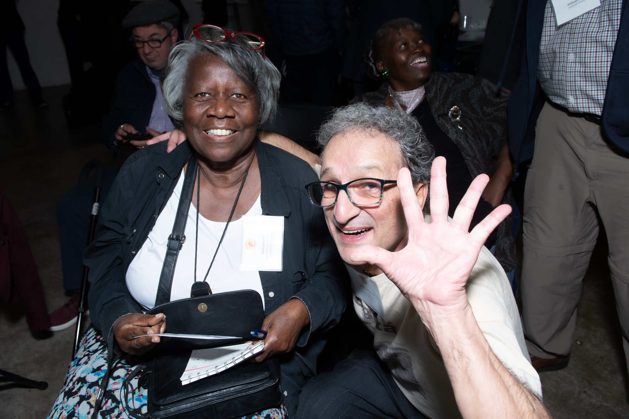 Two members of EBHO's Regional Community Organization Program pose for a photo at EBHO's 2019 Membership Meeting. The man sitting in front has five fingers raised to wave at the camera.