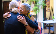 Former Board President Sister Marie Taylor faces the camera and hugs Vice President Vanna Whitney. She has a warm smile on her face.