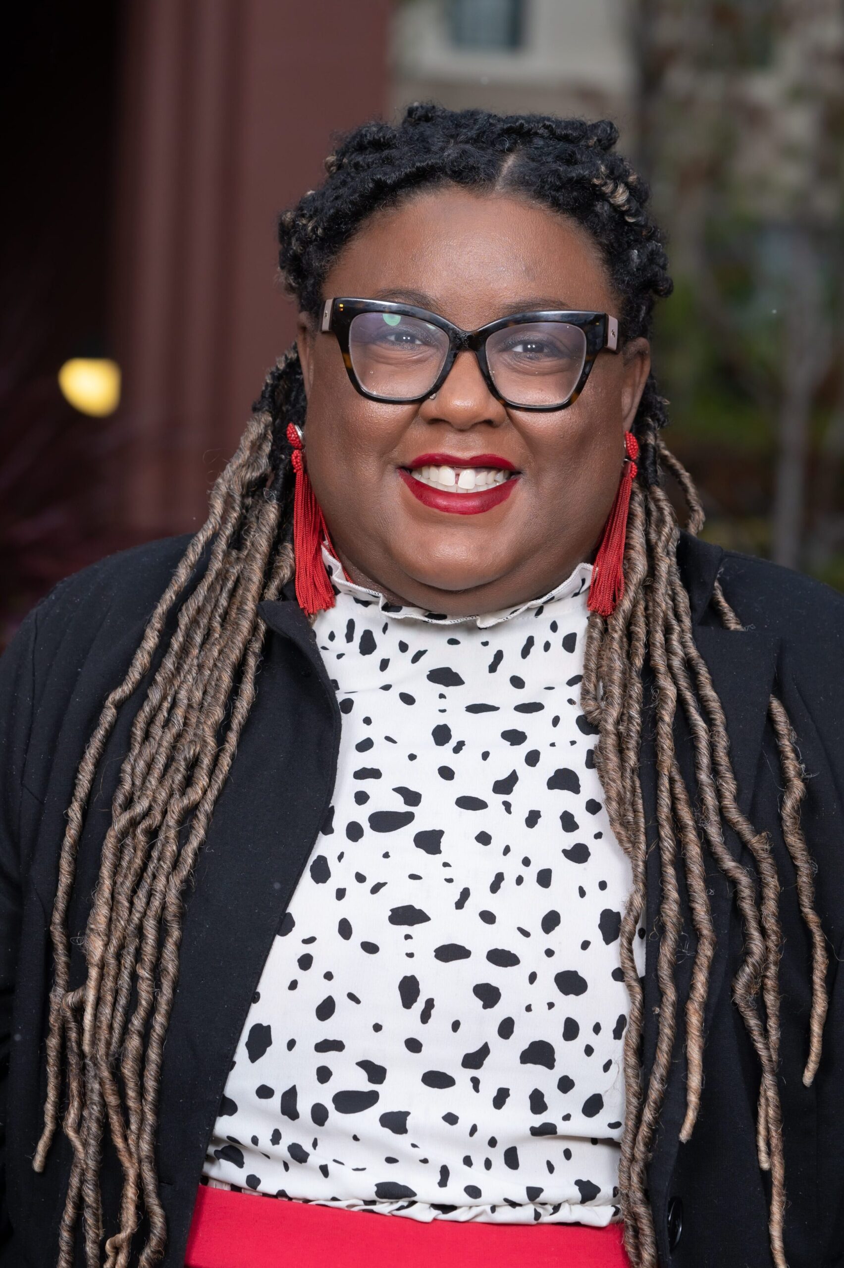 Ronnie Boyd smiles at the camera. She is a Black woman wearing thick-rimmed black glasses, bright red lipstick, and red earrings. Her hair is styled in braids.