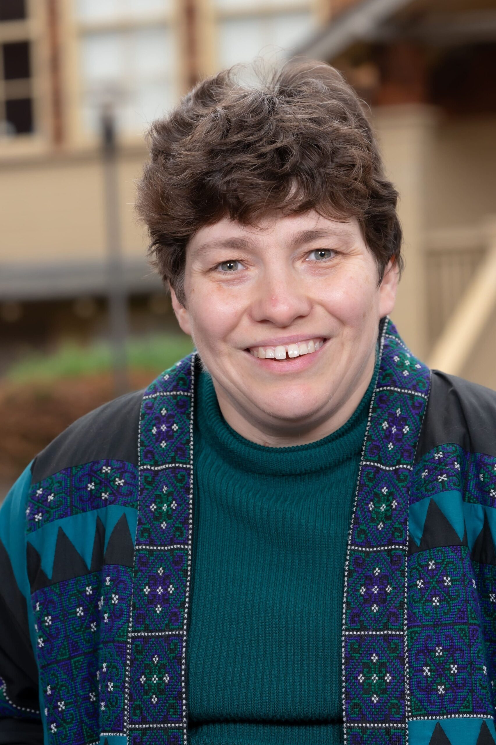Sophia DeWitt smiles at the camera. She is a white woman with short cropped, wavy hair. She is wearing a teal turtleneck and a patterned blue cardigan.