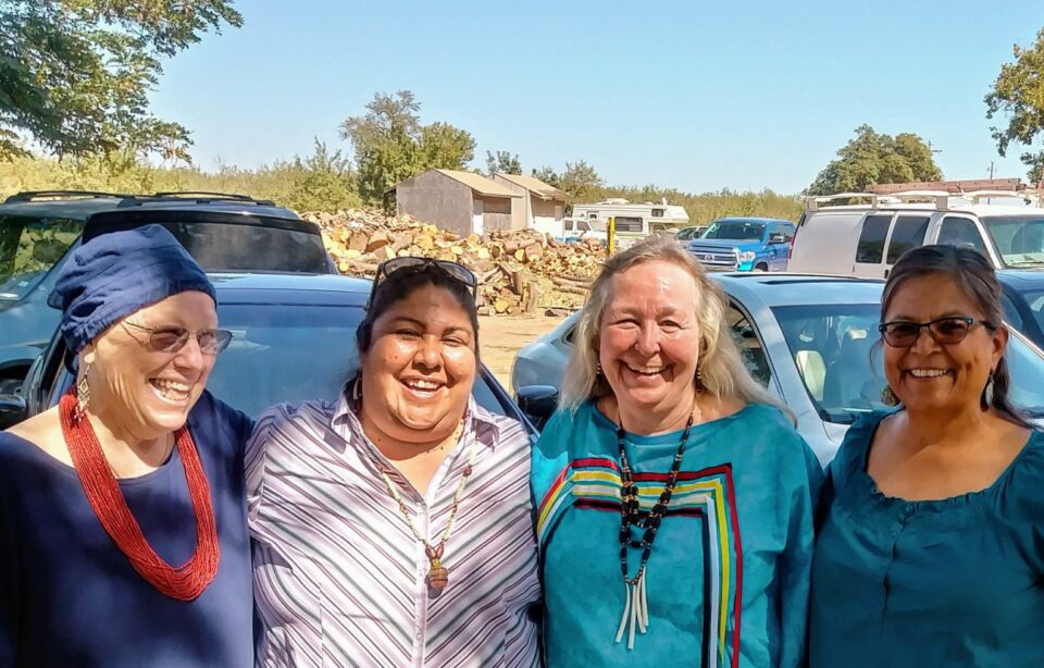 Four Indigenous women smile at the camera. They are of different generations and different tribes. The women wear traditional beaded jewelry as earrings and necklaces.