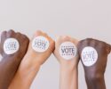 Four people with different skin tones raise their visits. Each of them has a "VOTE" sticker on the back of their hand.