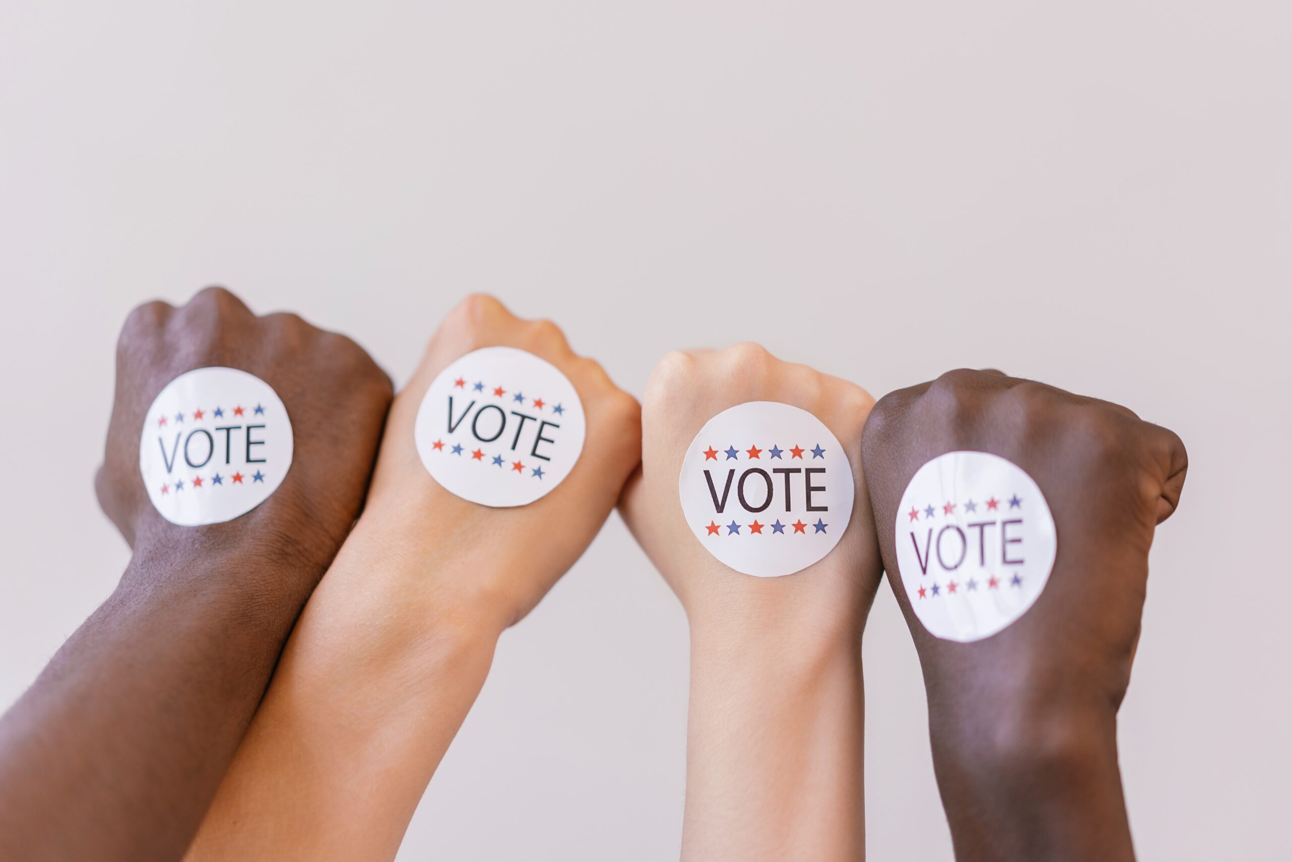 Four people with different skin tones raise their visits. Each of them has a "VOTE" sticker on the back of their hand.
