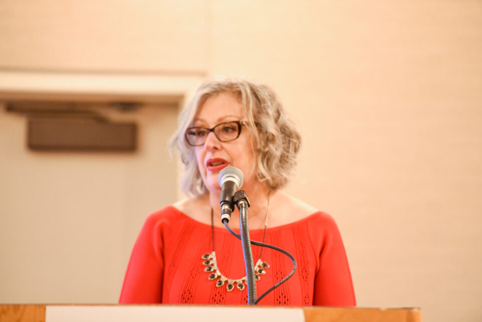 Debra Ballinger speaks into a microphone. She is a white woman with curly, shoulder-length hair wearing red lipstick and a red sweater.