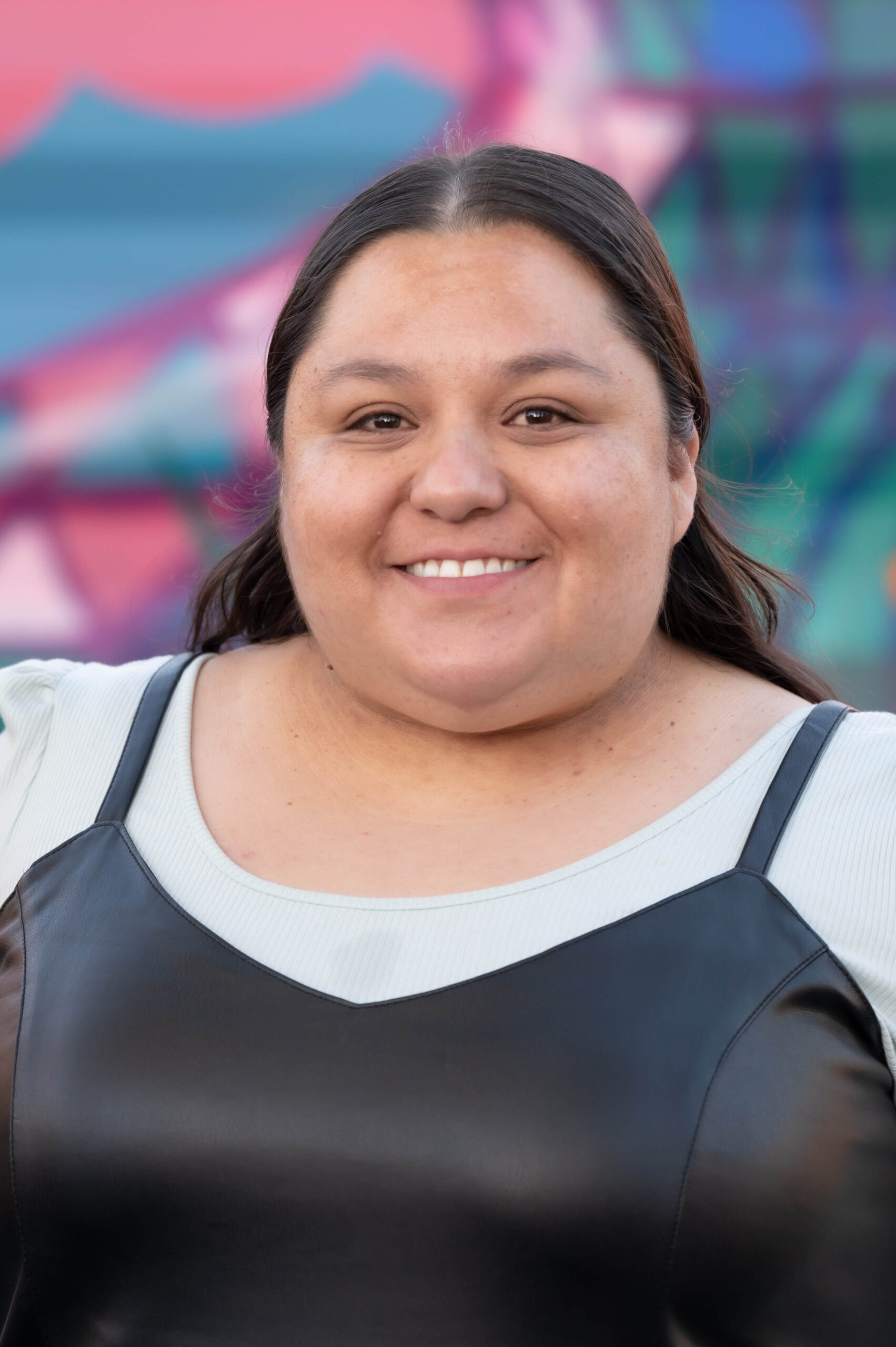 Dolores Tejada smiles for a headshot. They are a nonbinary person with their long brown hair pulled back off of their face. They are wearing a black strappy top over a white t-shirt.