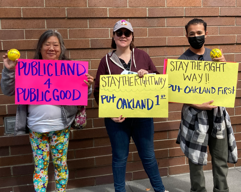 Picture of three protestors at a rally calling on the Oakland A's to come to the table and negotiate community benefits. Signs read "Stay the right way! Put Oakland first" and "Public land for public good"
