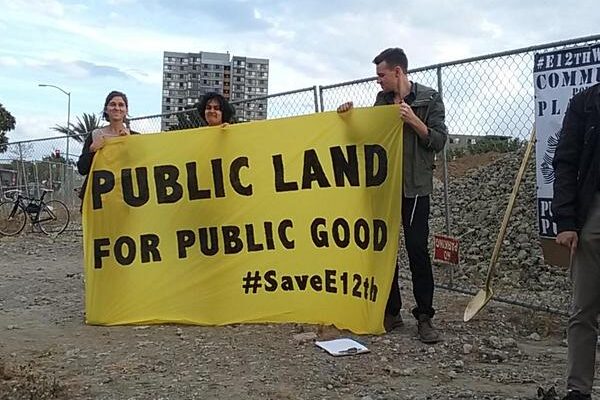 Three people stand on the vacant East 12th Street lot holding a large yellow banner that reads "Public Land for Public Good #SaveE12th"
